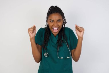 Sağlık üniforması giyen, yumruklarını sıkan, neşe ve heyecanla haykıran güçlü, kararlı genç doktor kadın portresi. Zafer, başarı ve başarı kavramı.