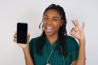 Heyecanlı Afrikalı Amerikalı doktor kadın akıllı telefon boş ekranı gösteriyor, göz kırpıyor ve el işareti yapıyor. Akıllı telefonu boş ekranla tutan şok olmuş bir kızın stüdyo fotoğrafı. Reklam kavramı.