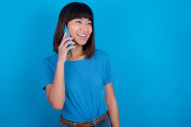 Mutlu görünümlü, mavi arka planda tişört giyen Asyalı genç bir kadın güzel bir telefon görüşmesi yapıyor ve yan gelip bakıyor, cep telefonuyla konuşurken güzel havasında ve gülümsüyor.