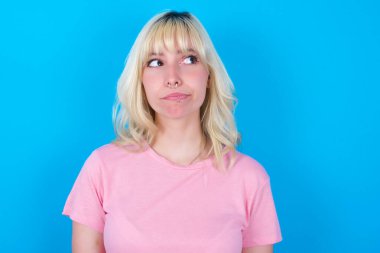 Şaşırmış Kafkasyalı kız pembe tişört giyiyor mavi arka plan kıvrımları üzerinde izole edilmiş dudaklar ve endişeli bir bakış, önünde korkunç bir şey görüyor.