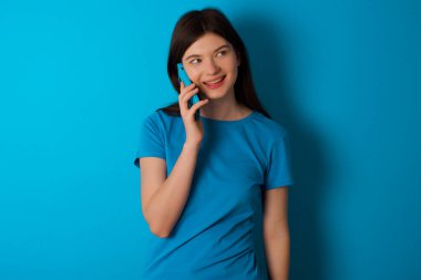 Mavi arka plan üzerinde izole edilmiş mavi tişört giyen mutlu görünümlü genç beyaz kız hoş bir telefon görüşmesi yapıyor ve yan tarafa bakıyor, cep telefonuyla konuşurken keyifli ve gülümsüyor.