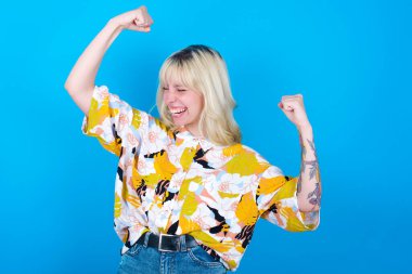 Evet, ben kazandım. Mavi arka planda izole edilmiş çiçekli gömlek giyen, mutlu ve heyecanlı beyaz bir kızın portresi zaferle ve zaferle gülümseyen, sevinçten sırıtan bir zafer gülümsemesiyle yumruğunu kaldırıyor..