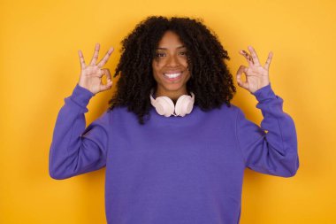 Sarı arka planda kulaklıklı Afro-Amerikan kadının portresi iyi jestler gösteriyor.