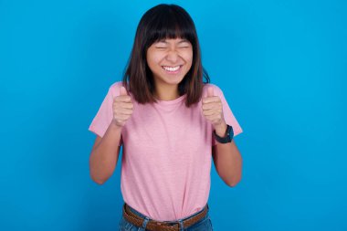 Mavi arka planda tişört giyen genç Asyalı kadın, onun başarısını ve zaferini sevinçle kutluyor. Amaç ve amaçlarına ulaştığına sevinerek yumruklarını sıkıyor. Olumlu duygular, duygular.