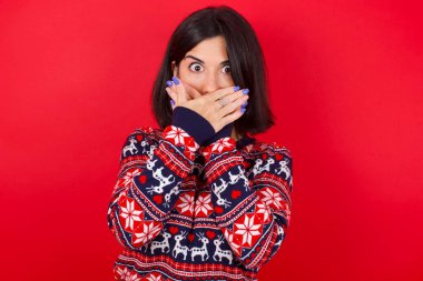 Kızıl arka planda Noel süveteri giyen esmer, beyaz kadın ağzını yanlışlıkla ellerle kapatmış. Gizli konsept.