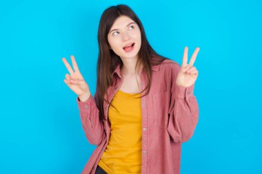 Mavi arka planda izole edilmiş pembe tişört giyen genç beyaz bir kızın izole edilmiş fotoğrafı iki elinle barış ya da zafer işareti yapar..