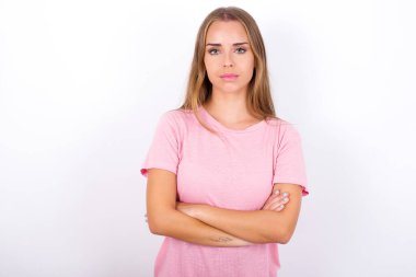 Kendine güveni tam, beyaz arka planda pembe tişört giyen, kolları bağlı, sakin, beyaz bir kız. Profesyonel titreşimlerin kendine güvenen pozlarda durduğunu gösterir.