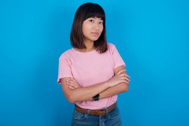 Mavi arka planda t-shirt giyen kendine güvenen genç Asyalı kadının belinde geniş bir gülümseme, kollarını kavuşturma, iş arkadaşlarıyla tanışmaktan mutlu..