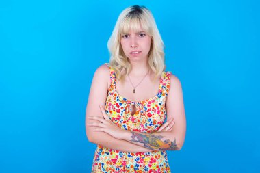 Mavi arka planda izole edilmiş çiçek desenli elbise giyen beyaz bir kız hoşnutsuzlukla yüzünü buruşturuyor, kollarını kavuşturup bir açıklama bekliyor..