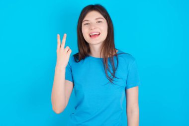Mavi tişört giyen genç beyaz kız mavi arka planda izole edilmiş gülümsüyor ve arkadaş canlısı görünüyor, iki ya da iki numara öne doğru, geriye doğru sayıyor.