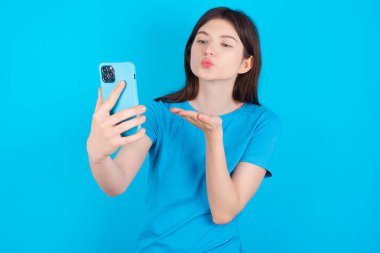 Mavi tişörtlü genç beyaz kız mavi arka planda izole edilmiş. Akıllı telefon kamerasına hava öpücüğü veriyor ve selfie çekiyor. İnternetten Mwah 'ı gönderiyor..