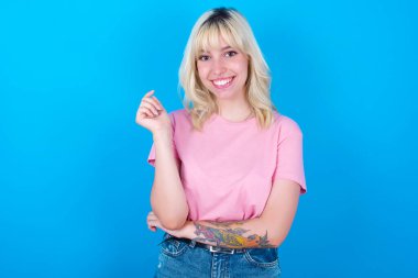 Pembe tişört giyen neşeli beyaz kız mavi arka planda izole edilmiş ve eli yüze yakın. İyi haberleri dinledikten sonra kameraya mutlu bir ifadeyle bakmak. Kendine güven..