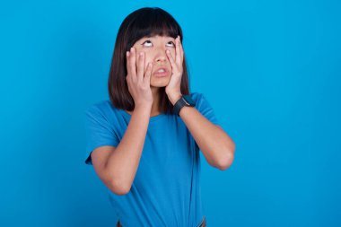 Mavi arka planda tişört giyen genç Asyalı kadın ellerini yanaklarından ayırmıyor ve sıkılmış bir ifade takınıyor. Stresli umutsuz model