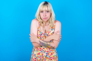 Mavi arka planda izole edilmiş çiçek desenli elbise giyen beyaz bir kız kış soğuğu yüzünden titriyor ve donuyor, yüzünde üzgün ve şok ifadesi var..