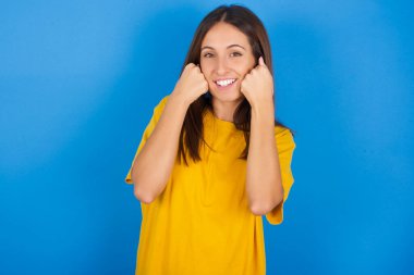 Mutlu genç kadın yumruklarını yanaklarına dayayıp gülümsüyor ve iyi bir ruh hali içinde pozitif bir ifade takınıyor.