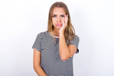 Beyaz arka planda çizgili tişört giyen üzgün, yalnız, güzel, sarışın kız yanağına dokunuyor, dudaklarını ısırıyor ve hoşnutsuzca bakıyor. Kötü duygular