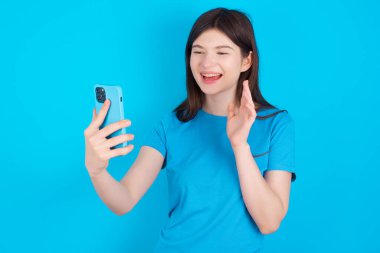 Mavi tişörtlü mutlu beyaz kızın portresi mavi arka planda izole edilmiş selfie çekiyor ve el sallıyor, video görüşmesinde iletişim kuruyor, internette sohbet ediyor..