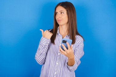 Güzel genç bir kadın başparmağıyla işaret ediyor ve boş alanı gösteriyor. Cep telefonunu mesaj göndermek için tutuyor..