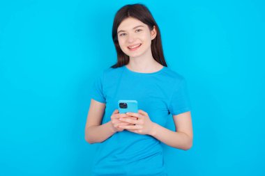 Mavi tişört giyen genç beyaz kız mavi arka planda izole edilmiş fotokopi alanı oluşturmuş. Cep telefonu kullanmak, SMS iletisi yazmak