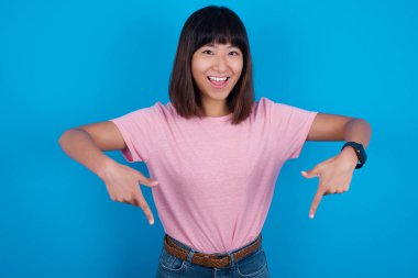 Mavi arka planda tişört giyen genç Asyalı kadın olumlu ifade ile işaret ediyor, her iki işaret parmağıyla, geniş ilgi çekici bir gülümsemeye sahip. Şuraya bakın, lütfen..