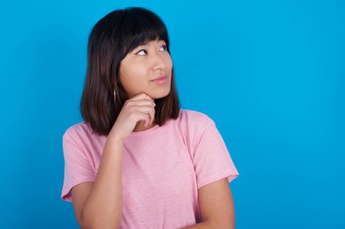 Yüz ifadeleri ve duygular. Düşünceli genç Asyalı kadın mavi arka planda tişört giyiyor. Başının altında el ele tutuşuyor, şüpheli bakıyor..