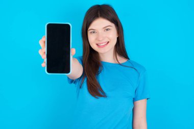 Mavi tişört giyen sevimli beyaz kız mavi arka planda izole edilmiş, elinde modern bir aletle siyah ekran akıllı telefon gösteriyor.