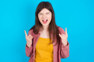 Bu dünyayı sallamak için doğmuşum. Mavi arka planda izole edilmiş pembe tişört giyen neşeli beyaz kız yüksek sesle bağırıyor ve kollarını kaldırarak ya da taş gibi hareketlerle gösteriyor..