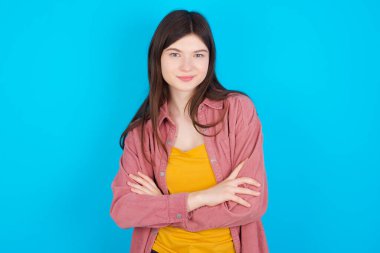 Kendine güvenen, mavi arka planda izole edilmiş pembe tişörtlü, kendine güvenen genç beyaz bir kızın belden yukarısında geniş bir gülümseme, kollar çapraz, iş arkadaşlarıyla tanıştığı için mutlu..