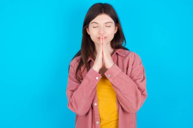 Mavi arka plan üzerinde izole edilmiş pembe tişörtlü genç beyaz kızın kapalı çekimleri. Yoga ve meditasyon yapıyor, namaste 'de avuç içlerini bir arada tutuyor, sakin, rahat ve huzurlu görünüyor..