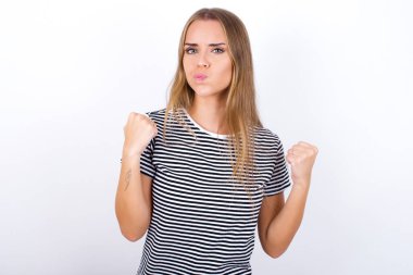 Beyaz arka planda çizgili tişört giyen sinirli sarışın kız öfkeyle yanaklarına vuruyor ve yumruklarını kaldırıyor öfke ve agresif duyguları ifade ediyor. Öfkeli model