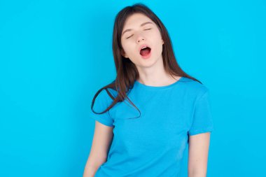 Mavi tişört giyen beyaz genç bir kız mavi arka plan esnemeleri ve ağzı açık esnemeler üzerine izole edilmiş. Günlük sabah rutini