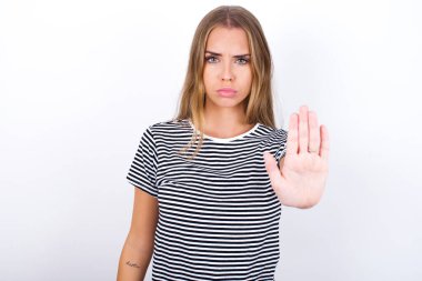 Beyaz arka planda çizgili tişört giyen güzel sarışın kız dur işareti gösteriyor. Avuç içini kameraya doğru sert bir ifadeyle çekiyor.