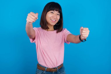 Mavi arka planda tişört giyen çekici Asyalı genç bir kadının portresi. Ellerini başının üstünde tutarak gülümsüyor. Güven ve rahatlama kavramı.