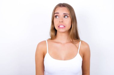 Tüh! Beyaz arka planda beyaz atlet giyen beyaz elbiseli genç beyaz kızın portresi dişlerini sıkar ve şaşırtıcı bir şekilde yana bakar.,