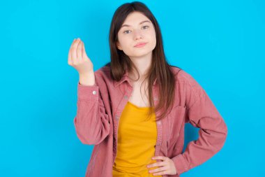 What the hell are you talking about. Shot of frustrated young caucasian girl wearing pink shirt isolated over blue background gesturing with raised hand doing Italian gesture, frowning, being displeased and confused with dumb question.