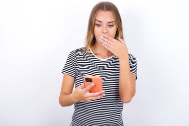 Beyaz arka planda çizgili tişört giyen güzel sarışın kız derinden şaşırıyor, akıllı telefon ekranına bakıyor, web sitesinde şok edici haberler okuyor, Omg, bu korkunç.!