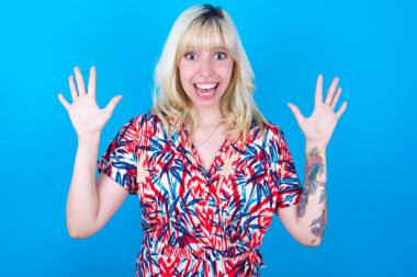 Mavi arka planda izole edilmiş çiçek desenli elbise giyen beyaz bir kız harika bir sonuç elde ettikten sonra ağzını ve kollarını açar.