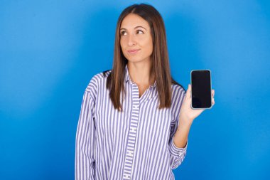 Güzel genç bir kadın yeni bir cep telefonu tutuyor ve gizemli görünüyor. Modern cep telefonunun boş görüntülerini gösteriyor.