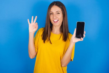 Heyecanlı genç bir kadın akıllı telefonuyla boş ekranı gösteriyor, göz kırpıyor ve el işareti yapıyor. Reklam kavramı.