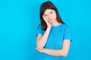 Mavi arka plan üzerinde izole edilmiş mavi tişört giyen üzgün, yalnız, genç, beyaz bir kız yanağına dokunuyor, dudaklarını ısırıyor ve hoşnutsuzca bakıyor. Kötü duygular