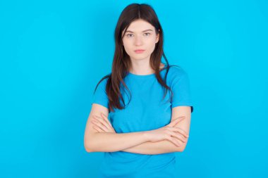 Mavi tişört giyen, kendine güveni tam genç beyaz bir kız. Mavi arka planda izole edilmiş, kolları çaprazlanmış, kameraya bakıyor.