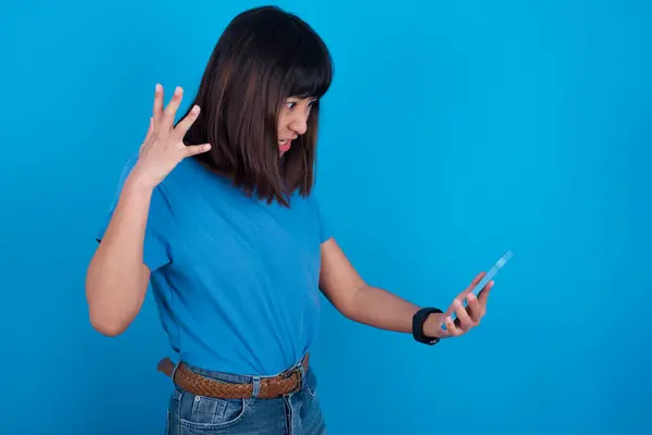 Photo of outraged annoyed young asian woman wearing t-shirt against blue background holds cell phone, makes call, argues with colleague,  expresses negative emotions. People and anger.