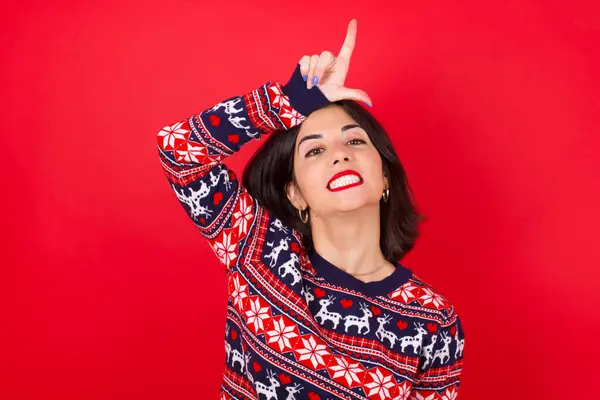 穿着红色背景的圣诞毛衣的女人取笑额头上有手指的人做失败者的手势嘲笑和侮辱 — 图库照片
