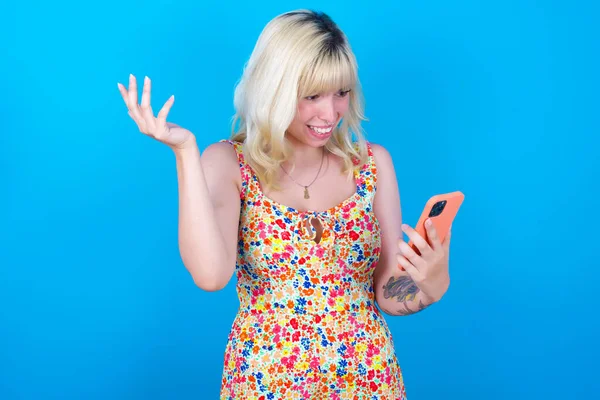 Photo of outraged annoyed caucasian girl wearing floral dress isolated over blue background holds cell phone, makes call, argues with colleague,  expresses negative emotions. People and anger.