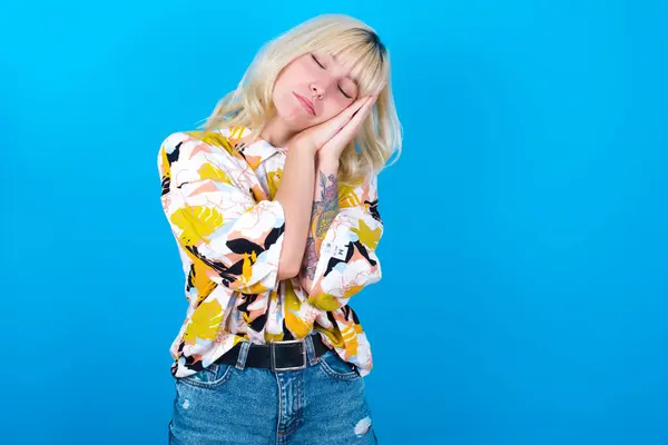 Relax and sleep time. Tired caucasian girl wearing floral shirt isolated over blue background with closed eyes leaning on palms making sleeping gesture.