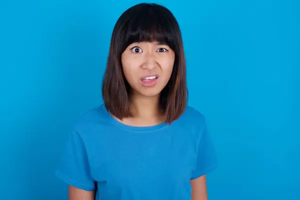 不满意的年轻亚洲女人的画像 穿着蓝色背景的T恤衫 面带微笑 嘴唇紧闭 恼怒地看着相机 不满地听到一些不愉快的声音 — 图库照片