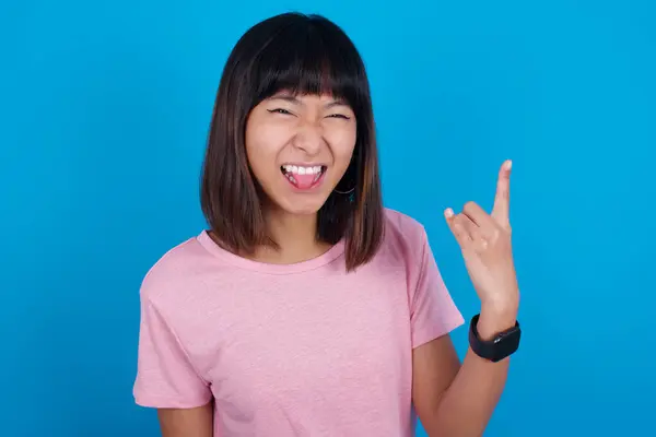 クレイジーな若いアジアの女性の肖像画 青い背景にTシャツを着て 舌の角がジェスチャーを示し バンドのコンサートにいることの興奮を表現 — ストック写真