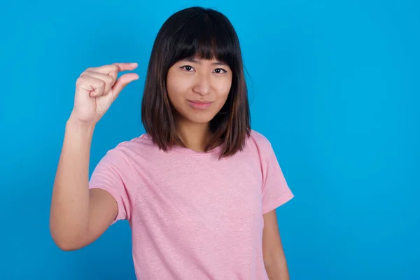 Upset young asian woman wearing t-shirt against blue background shapes little gesture with hand demonstrates something very tiny small size. Not very much