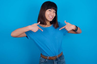 Beni seç! Kendine güveni tam, karizmatik, mavi arka planda tişört giyen genç Asyalı kadın kendini geniş gülümsemeli ve vücudu işaret eden bir rol istiyor olarak tanıtıyor..