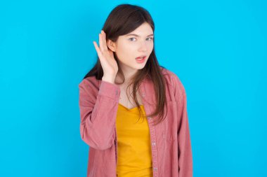 - Aman Tanrım! Mavi arka plan üzerinde izole edilmiş pembe tişört giyen komik, genç, beyaz bir kız ağzı açık bir şekilde yan tarafa bakıyor, elleri kulağına yakın, dedikoduları dinlemeye çalışıyor..